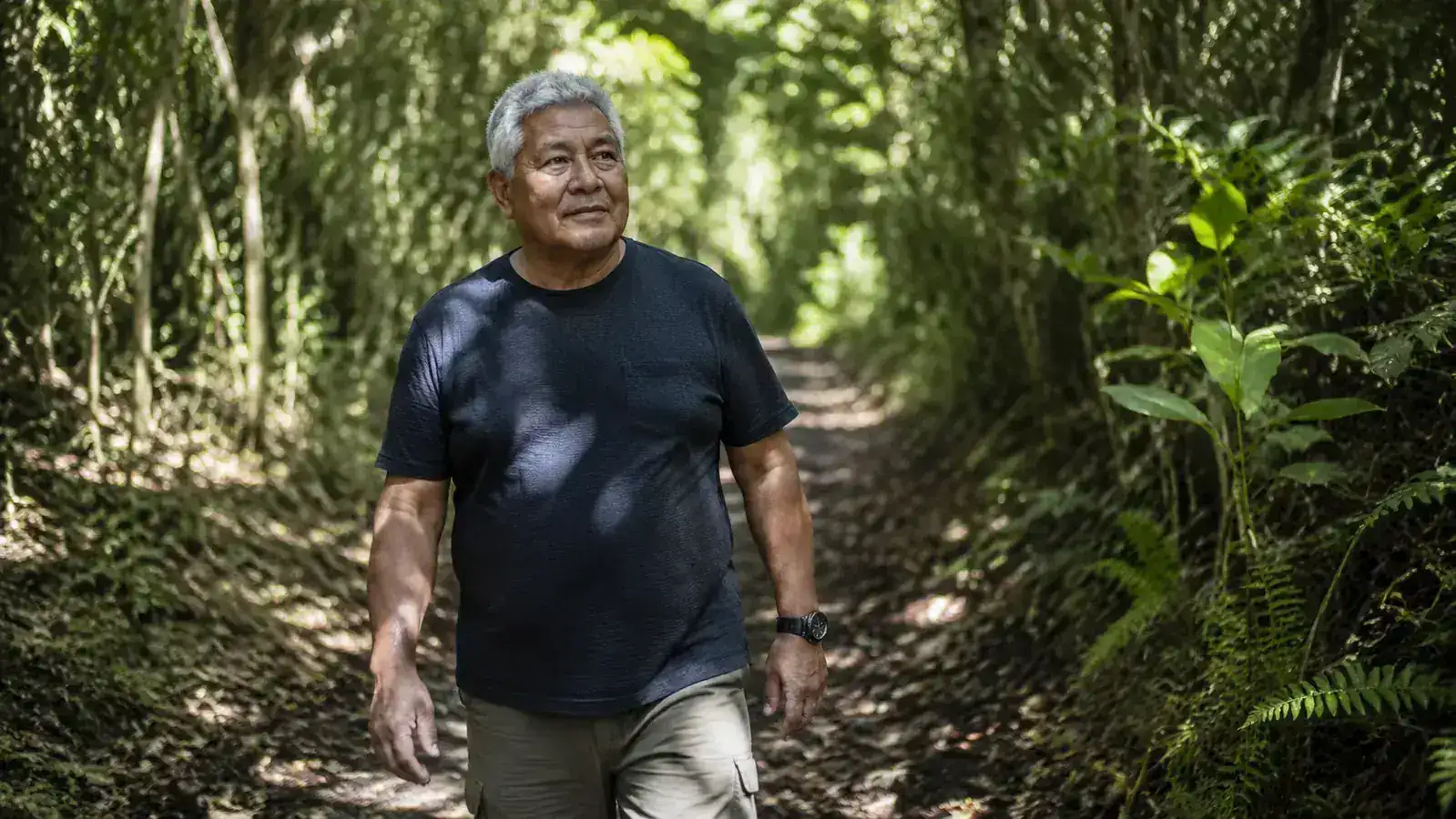 Older man walking a shaded forest trail, dappled light through canopy, looking alert and present